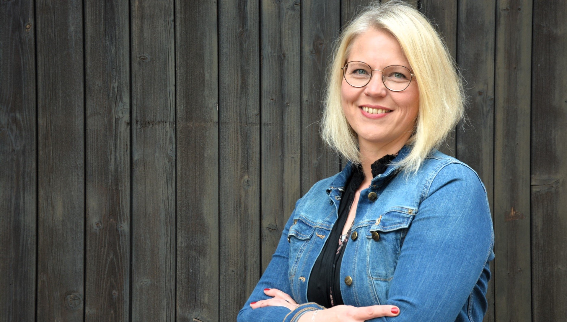 Tina Niemann steht vor einem dunklen Scheunentor, lachend, mit verschränkte Armen.
