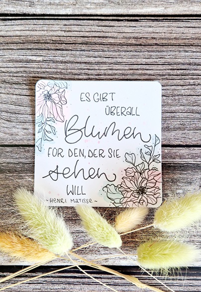 Zu sehen ist ein quadratisches Handlettering mit dem Text "Es gibt überall Blumen für den, der sie sehen will". Es handelt sich um ein Zitat von Henri Matisse. Das Lettering ist umrandet von Blumen-Ranken. Unter dem Lettering liegt eine Art Weidekätzchen-Deko, gelb eingfärbt.