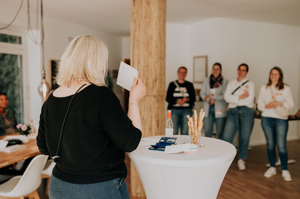 Tina steht mit dem Rücken zur Kamera, hält ein visuelles Symbol hoch, während sie ihren Vortrag hält. Im Hintergrund, unscharf, stehen Teilnehmerinnen, die aufmerksam teilhaben.