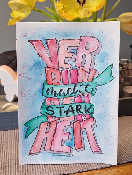 Im Vordergrund ein Lettering in Pink-Tönen und Grün mit den Worten "Verbundenheit macht stark". Im Hintergrund gelbe Tulpen und ein Porzellan-Schmetterling.