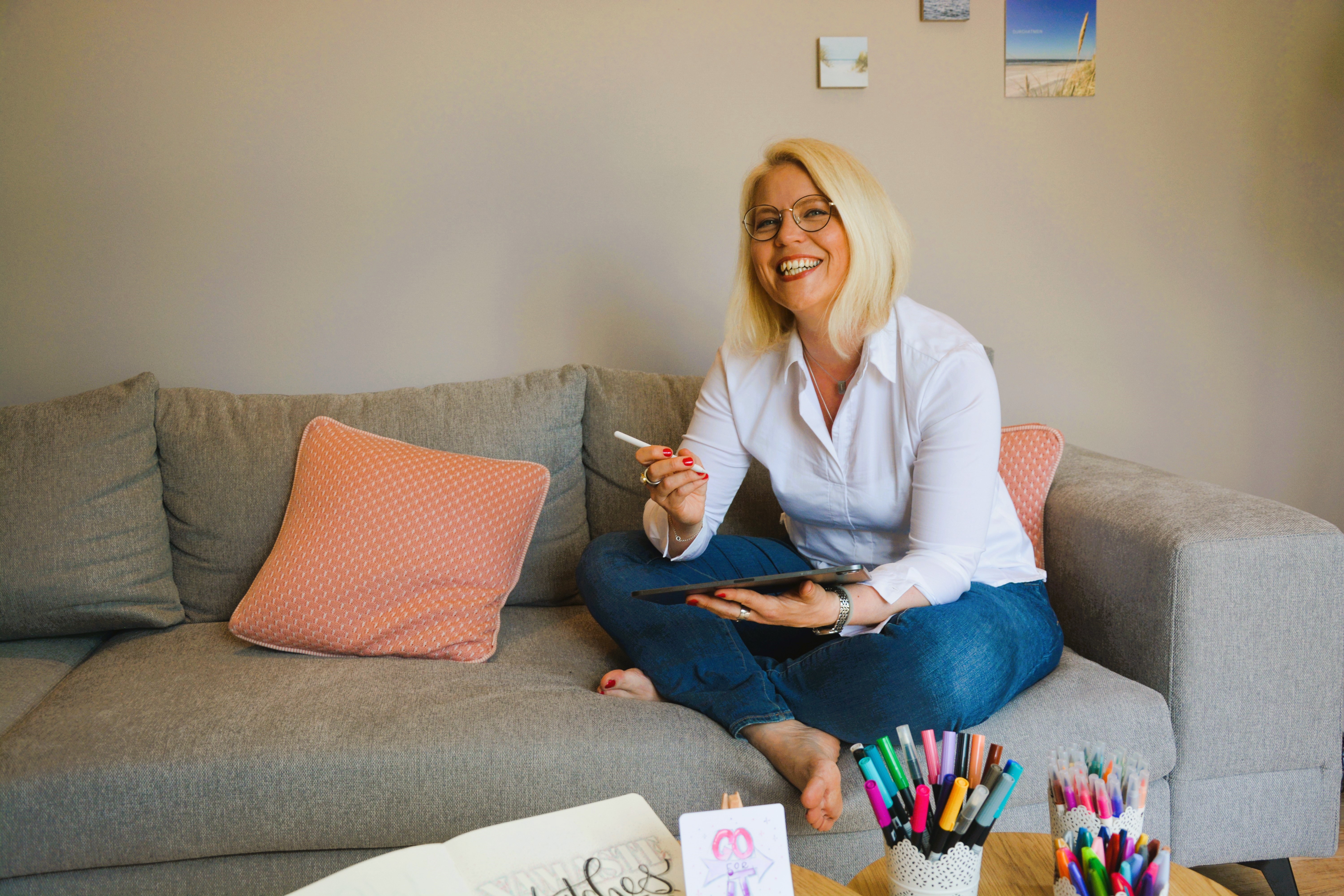 Tina sitzt auf der Couch mit ihrem IPad in der Hand. Vor ihr auf dem Tisch eine bunte Auswahl an Stiften.