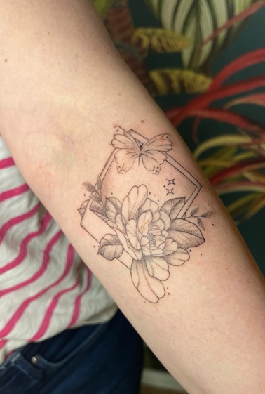 Tinas Unterarm mit ihrem Tattoo: Ein Polaroid mit einem Schmetterling, kleinen Sternen und einer geöffneten Pfingstrose, die aus dem Polaroid hinaus ragt.