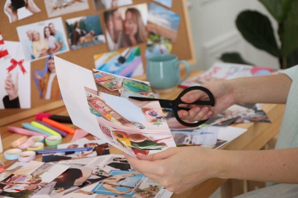 Stockfoto aus Canva: Ein Visionboard erstellen - Im Bild ein kreativer, chaotischer Schreibtisch mit viel Bastelmaterial. Im Vordergrund zwei Hände, die ein Blatt und eine Schere halten, eine Bild zurechtschneiden.