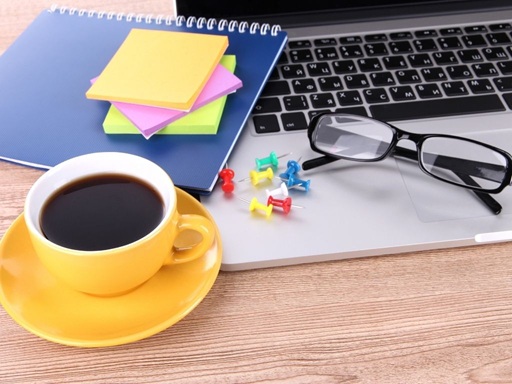 Ein chaotischer Tisch mit einem Laptop, kaltem Kaffee, einem Notizbuch, einer Lesebrille und ein paar Stecknadeln