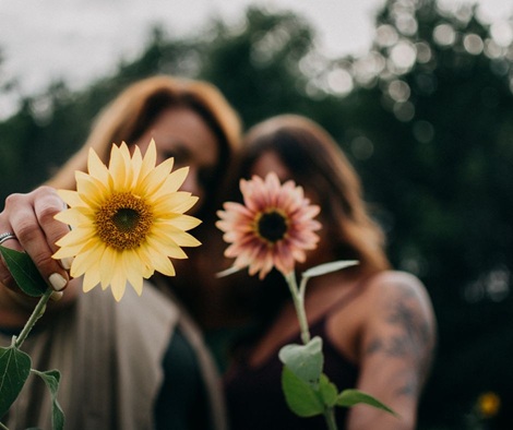 Im Hintergrund, verschwommen, sind zwei Frauen zu erahnen, die freundschaftlich ihre Köpfe aneinander lehnen. Im Vordergrund, schaft gestellt, hält jede von ihnen eine Blume in die Kamera. Links mit gelber Blüte, rechts mit roter Blüte.