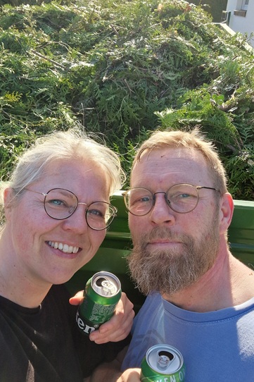 Tina und ihr Partner vor einem Anhänger voll Hecken-Schnitt, mit einer Dose Bier als Belohnung in der Hand