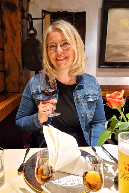 Tina sitzt im Restaurant am gedeckten Tisch, in der Hand ein Glas Wein