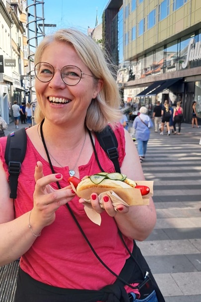 Tina in einer belebten Innenstadt mit einem dänischen Pölser in der Hand und einem strahlenden Gesicht